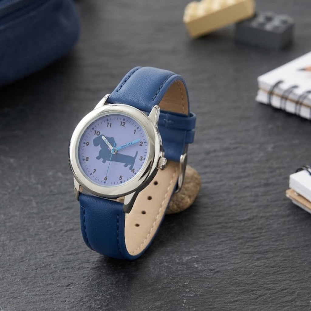 A wristwatch with a blue leather strap and a light blue dial featuring a dachshund silhouette, displayed on a dark slate surface with school-themed props like notebooks, a pencil, and toy blocks.