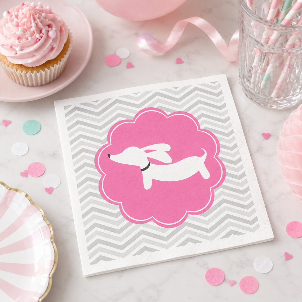 A square white napkin with a gray chevron pattern and a pink cloud-shaped graphic containing a white dachshund silhouette, styled on a marble surface with pink party props including a cupcake, candy jar, and confetti.
