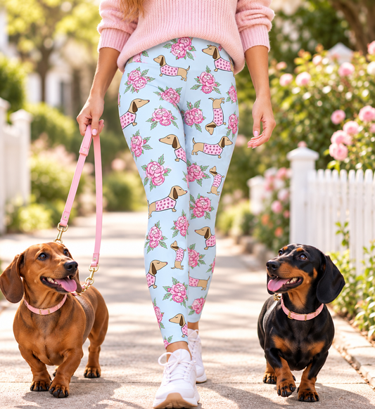 A person walking two dachshunds on a sunny outdoor path, wearing light blue leggings with a pink and brown dachshund and rose print, a pink sweater, and white sneakers.