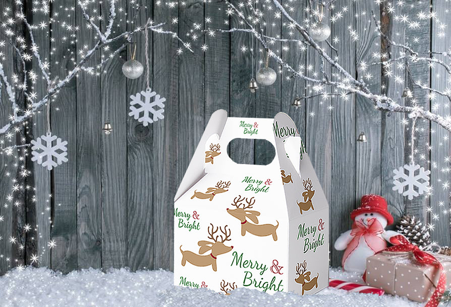 A white Christmas gift box with handle, decorated with brown reindeer illustrations and green 'Merry & Bright' text, set against a rustic gray wooden background with festive lights, snowflakes, and a small snowman decoration.