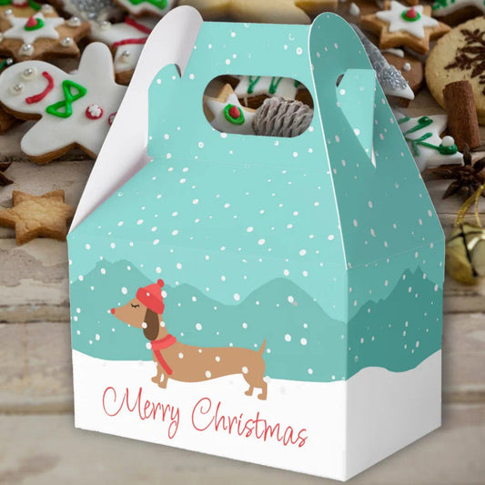 A Christmas-themed gift box with a handle, featuring a light blue winter scene with snowflakes, a dachshund wearing a red hat and scarf, and 'Merry Christmas' printed on the front. The box is shown filled with cookies and surrounded by holiday decorations on a wooden surface.